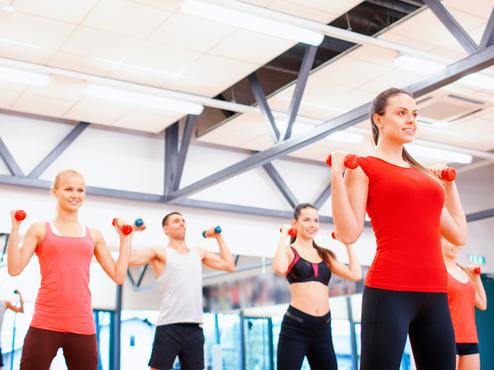 Gruppe von Menschen in Sportkleidung hebt Hanteln in einem Fitnessstudio mit Spiegeln und heller Beleuchtung.